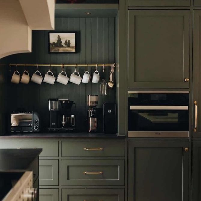 A dark green, modern kitchen featuring a built-in microwave, espresso machine, and a row of hanging mugs.