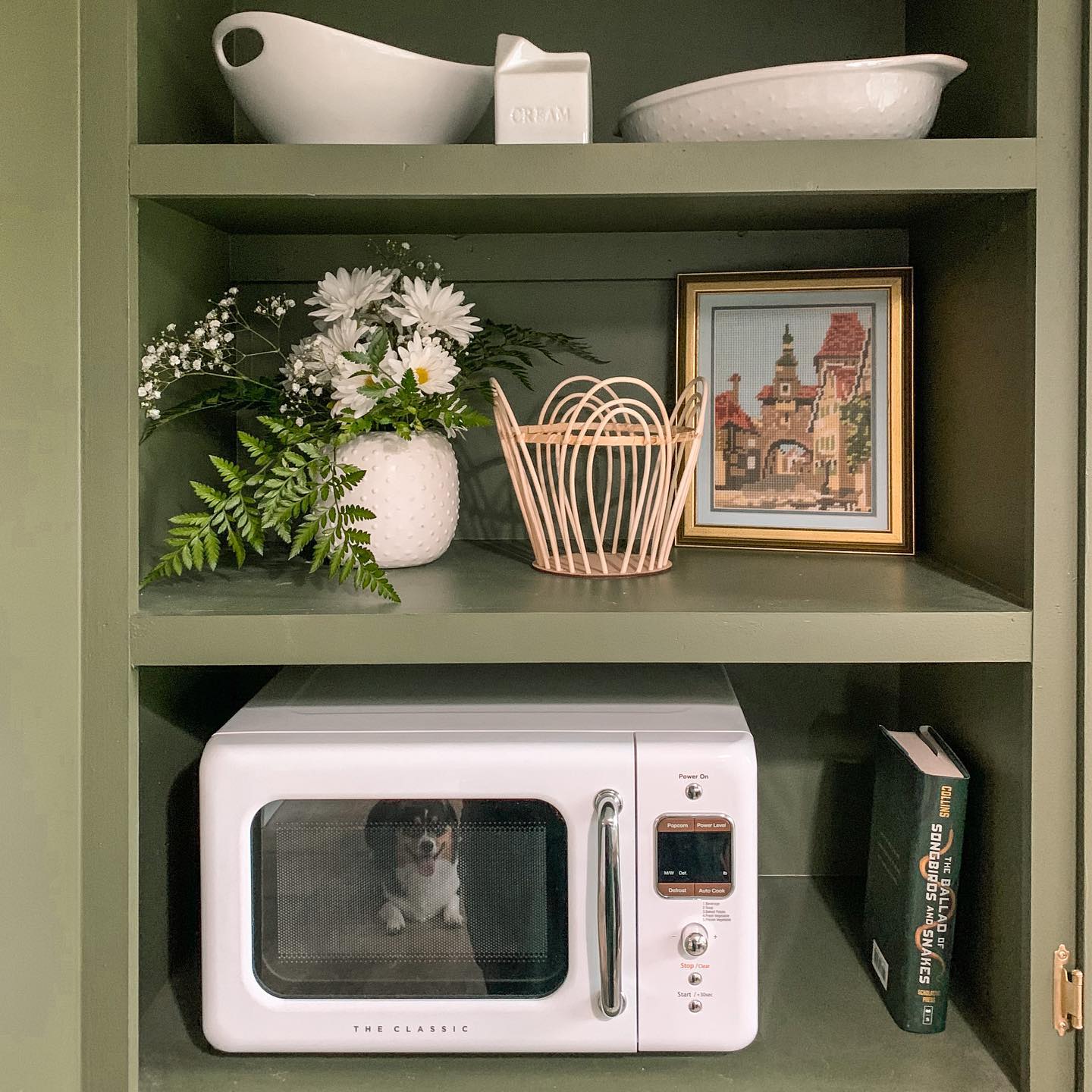 A white retro microwave sits on a green shelf, surrounded by various decorative items including a framed picture, flowers, and books.