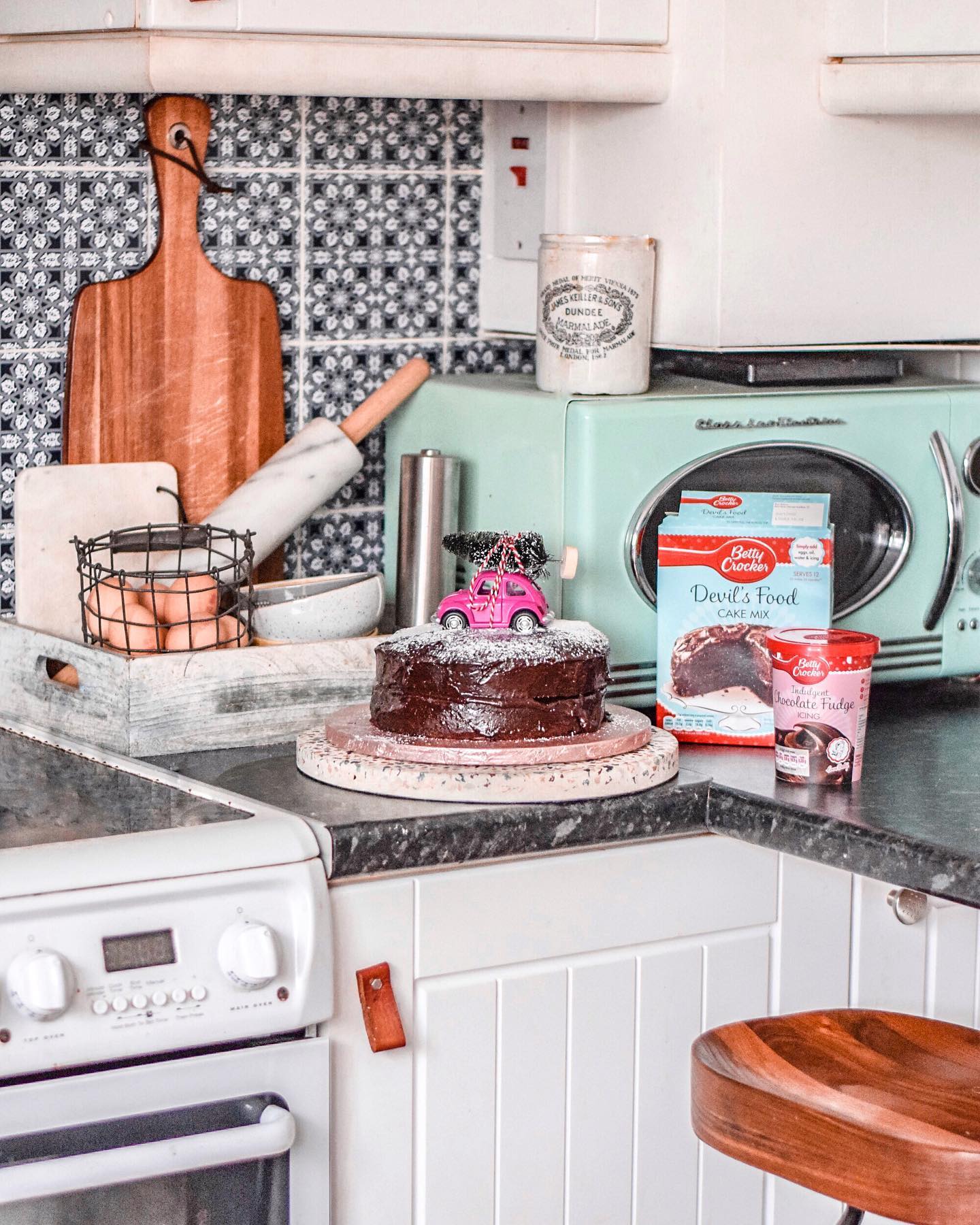 A retro-style kitchen with a teal microwave, a chocolate cake with a pink toy car on top, and various baking items.