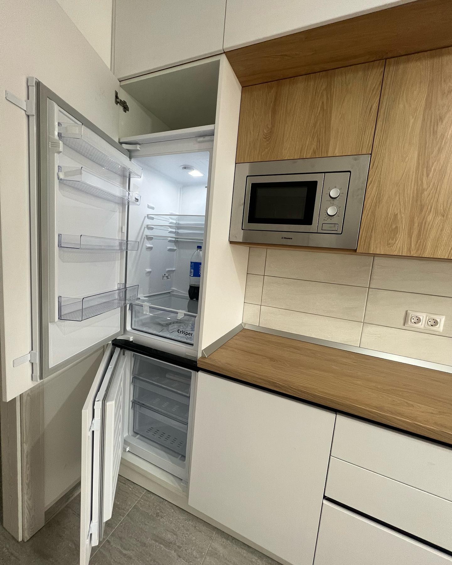 An image of a modern kitchen featuring an open fridge and a built-in microwave oven installed in wooden cabinetry above a white countertop.