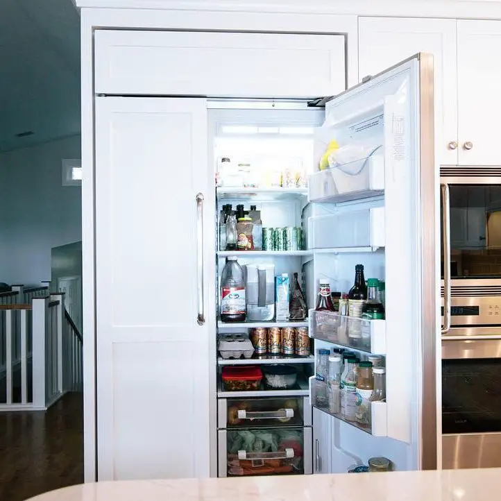 An open panel ready refrigerator filled with various food items, integrated seamlessly with the white cabinetry in a modern kitchen.