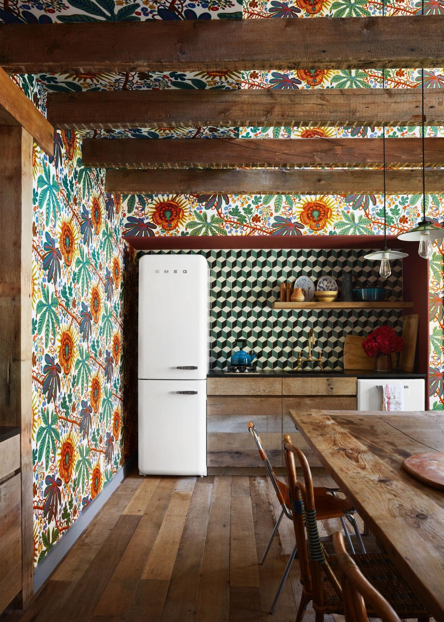 A white retro-style refrigerator stands against a wall adorned with colorful floral wallpaper, next to a wooden dining table and chairs.