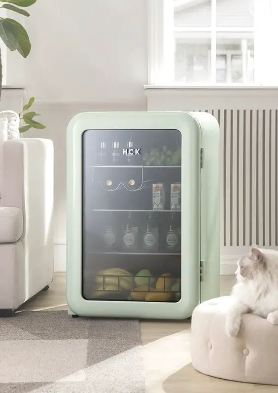 A mint green, vintage-style compact refrigerator with a transparent door, revealing beverages and snacks inside, placed in a bright living room. 