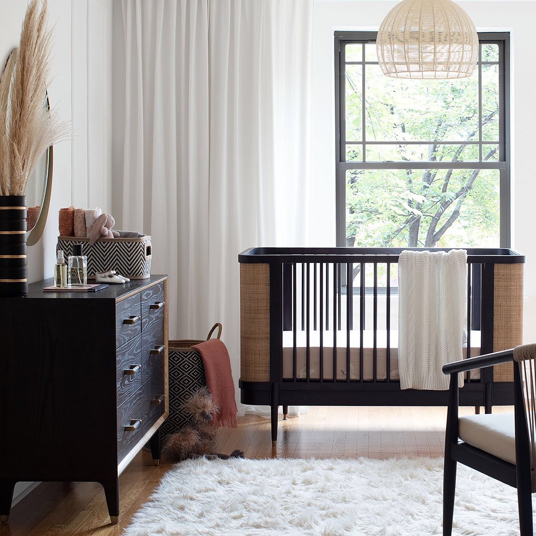 A bright and airy nursery room with a dark wood crib, a dresser, and a fluffy white rug. A large window lets in natural light and shows greenery outside.