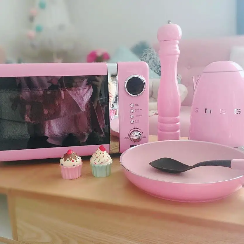 A pink retro microwave surrounded by other pink kitchen items including a pepper mill, a kettle, two cupcakes, and a frying pan with a spatula.