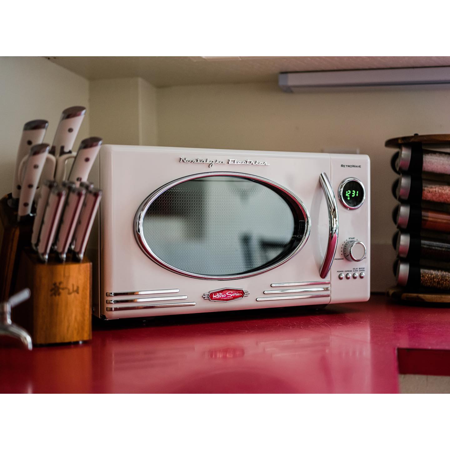 A white retro-style microwave with chrome detailing, digital display, and a round window, placed on a red countertop.