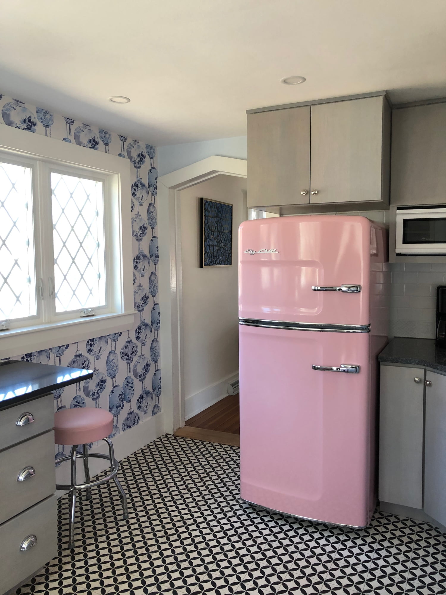 A pink retro-style refrigerator in a kitchen with blue floral wallpaper, black and white checkered floor, and modern cabinetry.