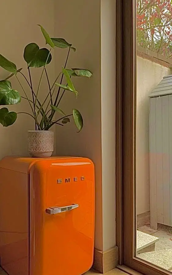 An orange SMEG refrigerator sits beside a window, with a potted plant on top.