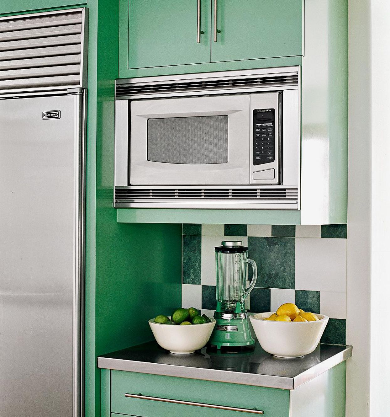 A built-in microwave oven installed above a countertop in a modern kitchen, with a blender and two bowls of fruits on the counter. 