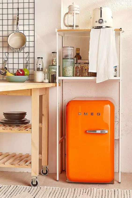 An orange SMEG refrigerator is placed under a white shelving unit, surrounded by various kitchen items including a coffee maker, fruits, and utensils. 