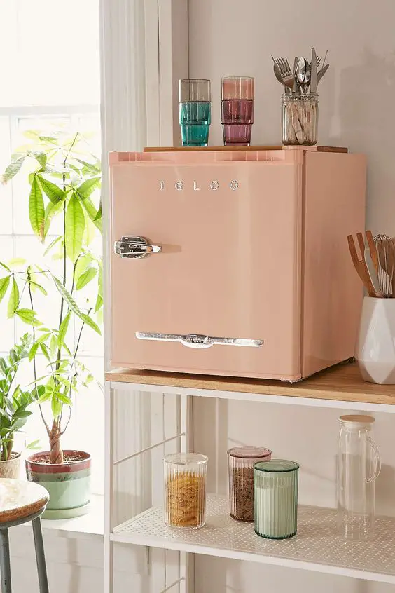  A small pink refrigerator with the brand name “IGLOO” on the front, placed on a wooden shelf beside a window, with various kitchen items around it. 