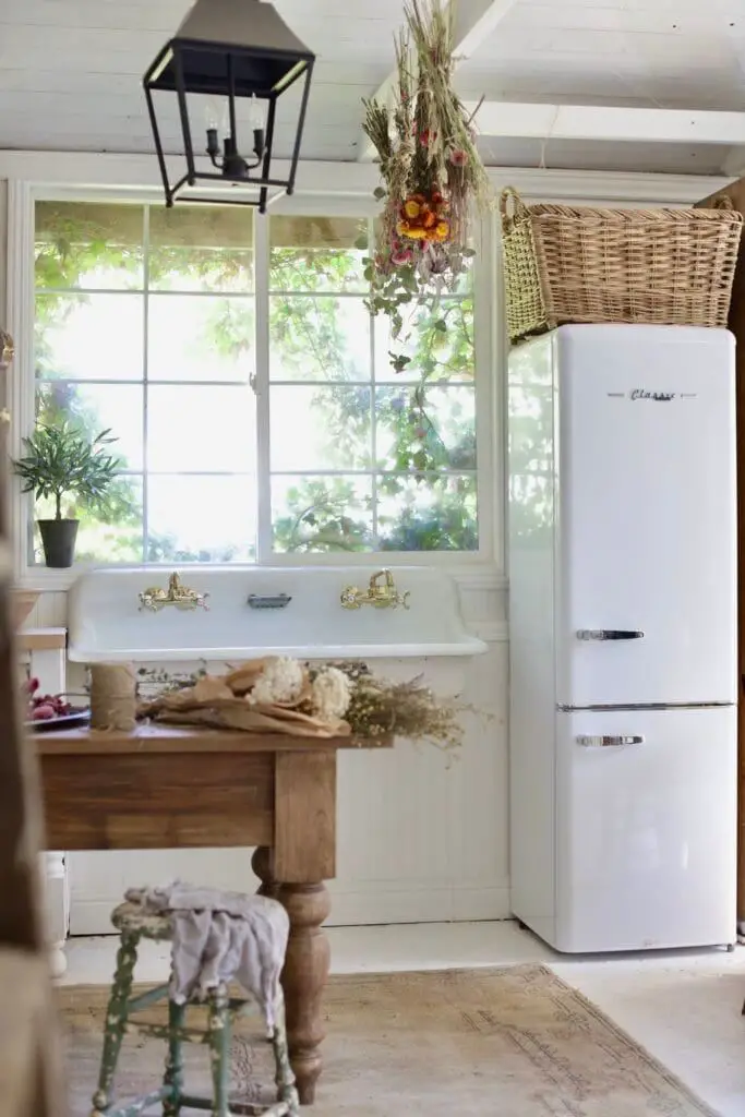 A white retro refrigerator beside a classic farmhouse sink, under a window with greenery outside, creating an atmosphere of vintage elegance.