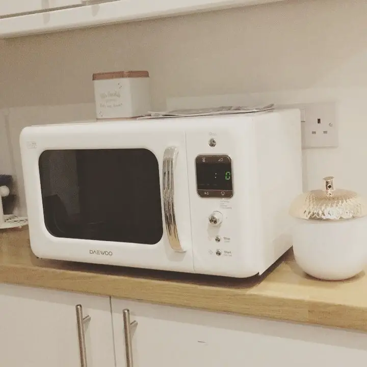 A white retro-style DAEWOO microwave with silver accents, digital display, and control knobs, placed on a wooden shelf in a light-toned modern kitchen.