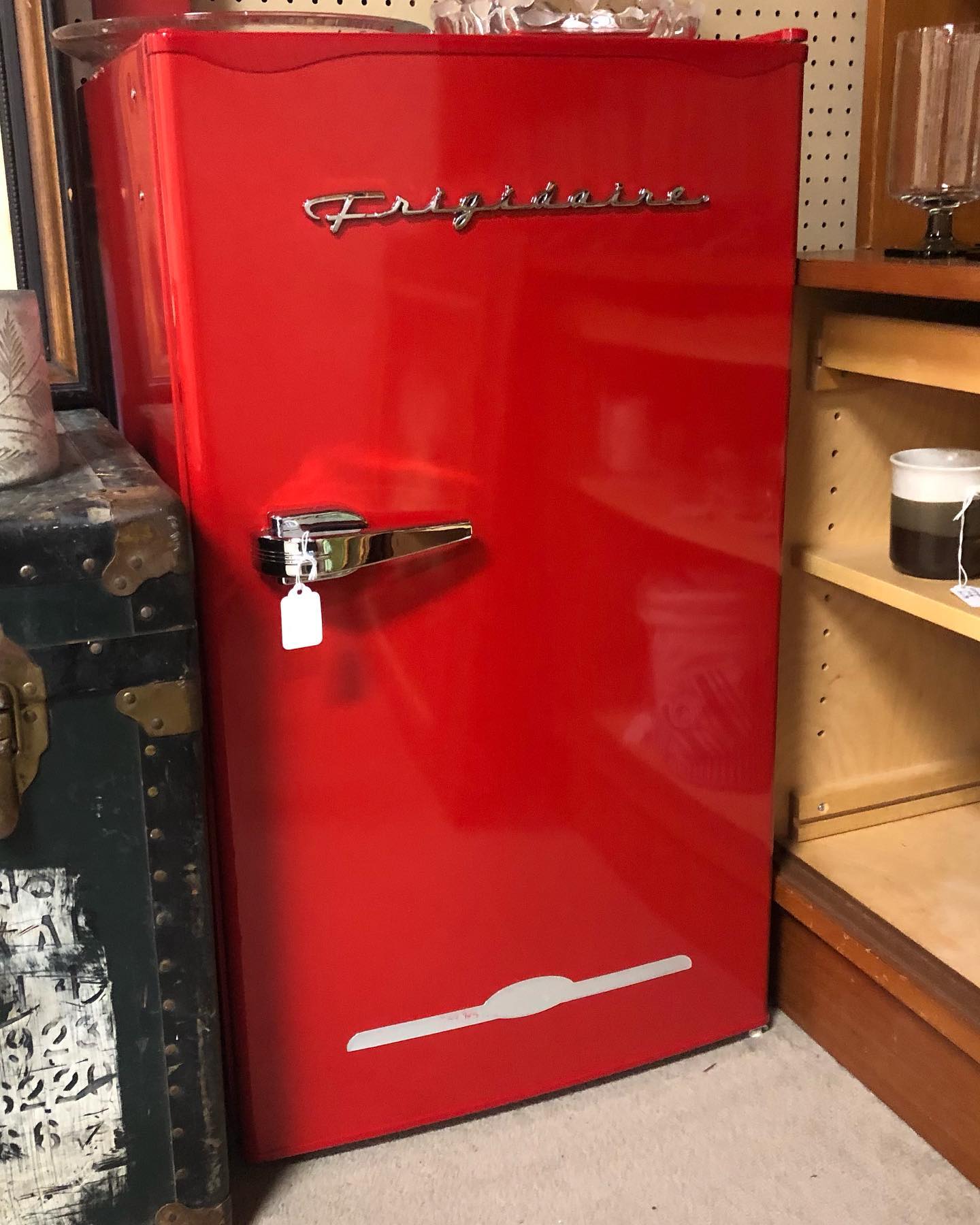 A bright red vintage Frigidaire refrigerator with a chrome handle, situated next to a wooden shelf and an old trunk.