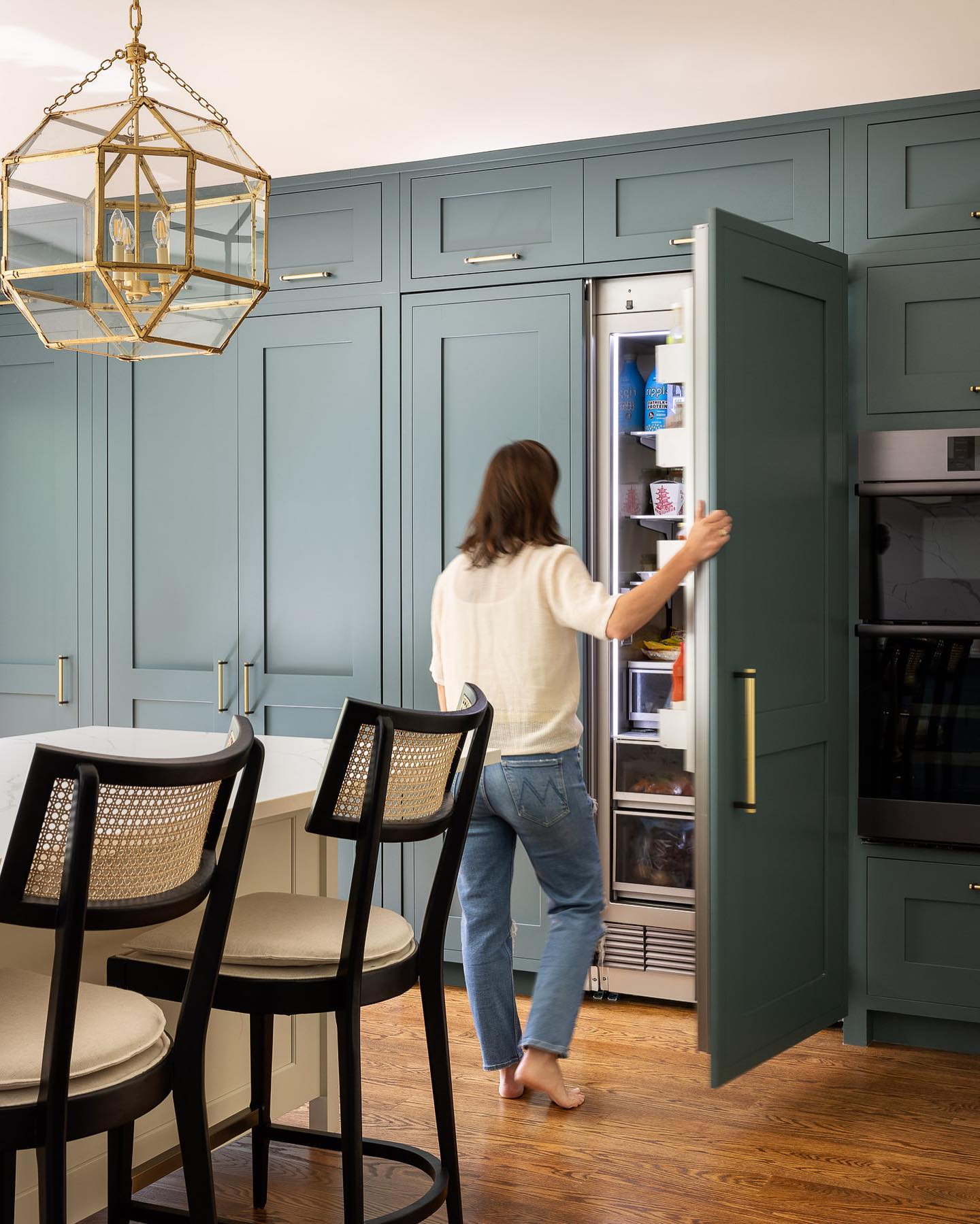 A person opening a panel-ready refrigerator integrated into teal cabinetry, with bar stools and a decorative light fixture in the foreground.